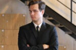 A person in a suit with arms crossed stands in front of a staircase, looking to the side. The background has wooden panels and metal railings.