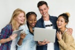 A group of four people smiling and looking at a laptop and tablet together, representing collaboration and technology use.
