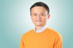 A headshot of a person with short black hair, wearing an orange sweater over a light blue collared shirt, smiling against a light blue background.