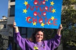 A person holding a blue sign with yellow stars and red letters reading "Our Future Our Choice" at a protest or rally. Buildings and trees are in the background.