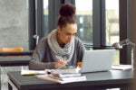 A person studying at a desk with a laptop, writing in a notebook, surrounded by books, and seated near large windows.