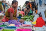 A person sitting at a market stall surrounded by vibrant colored powders, likely used for festivals like Holi, and fresh green leaves. Other market-goers are visible in the background.