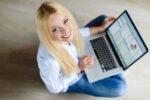 Person smiling while working on a laptop with charts on the screen, sitting on a light wood floor, viewed from above.