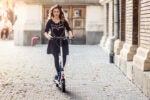 A person riding an electric scooter down a cobblestone street in an urban setting. The individual is wearing a black outfit and has long hair. The scene is well-lit with a warm, sunny ambiance.