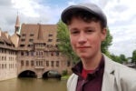 A smiling person in a flat cap and light jacket stands near a historical building by a river. The building has medieval architecture with a pointed spire.