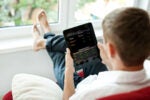A person relaxing near a window, using a tablet displaying a stock trading app with financial data and stock performance metrics.