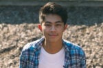 A person outdoors wearing a blue plaid shirt over a white T-shirt, smiling at the camera with a blurred background.