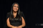 A person with long dark hair is smiling and sitting against a black background. They are wearing a black outfit and jewelry.
