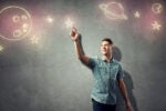 A person in a checkered shirt stands against a gray wall, pointing at chalk-like drawings of planets and stars, symbolizing imagination or aspiration.