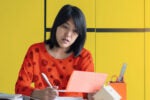 A person in a red polka dot top is reviewing a document at a desk. They are surrounded by stationery, with a yellow background.