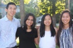 A group of four people smiling outdoors, standing closely together with trees and a house in the background.