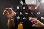 Hands interacting with a virtual interface displaying various icons related to the Internet of Things (IoT), such as locks, graphs, cars, and drones.