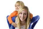A smiling adult with long blonde hair giving a piggyback ride to a child in an orange shirt. The background is plain white.
