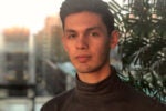 A person with short brown hair wearing a dark turtleneck sweater, standing indoors near a plant, with a blurred cityscape in the background.