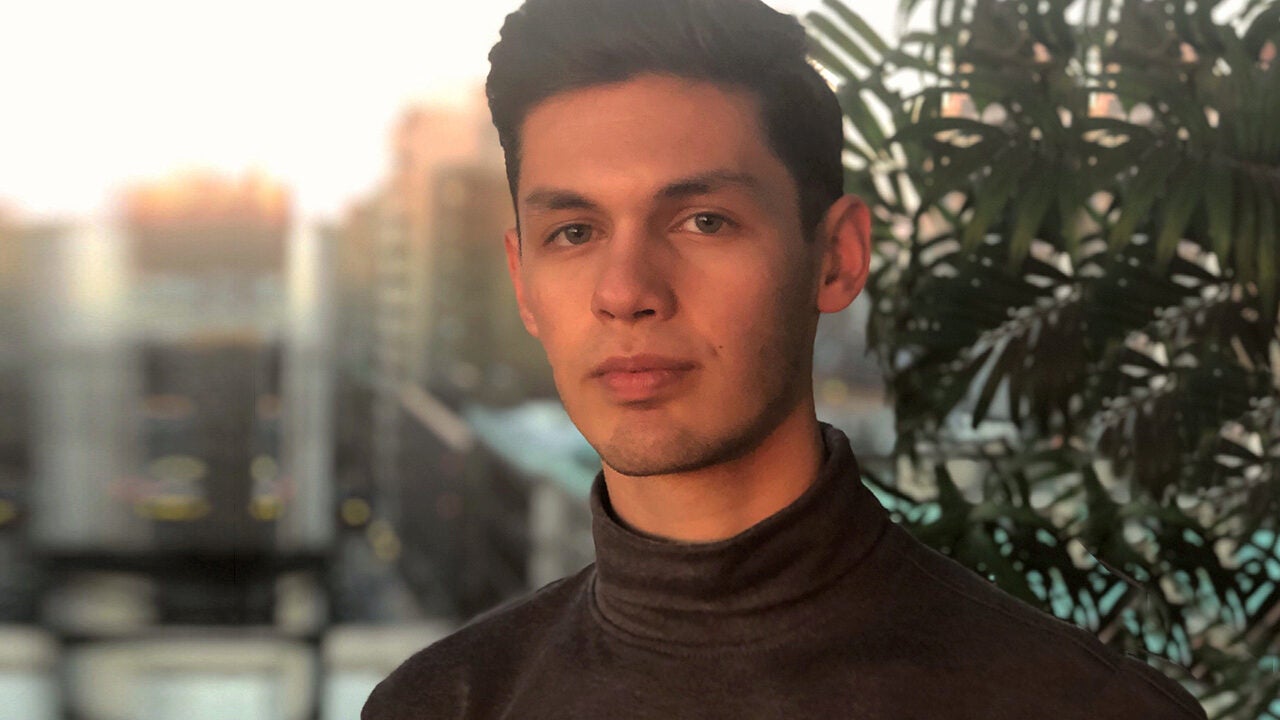 A person with short brown hair wearing a dark turtleneck sweater, standing indoors near a plant, with a blurred cityscape in the background.