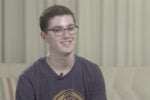 A person with short dark hair, wearing glasses and a t-shirt, sitting on a sofa and smiling.