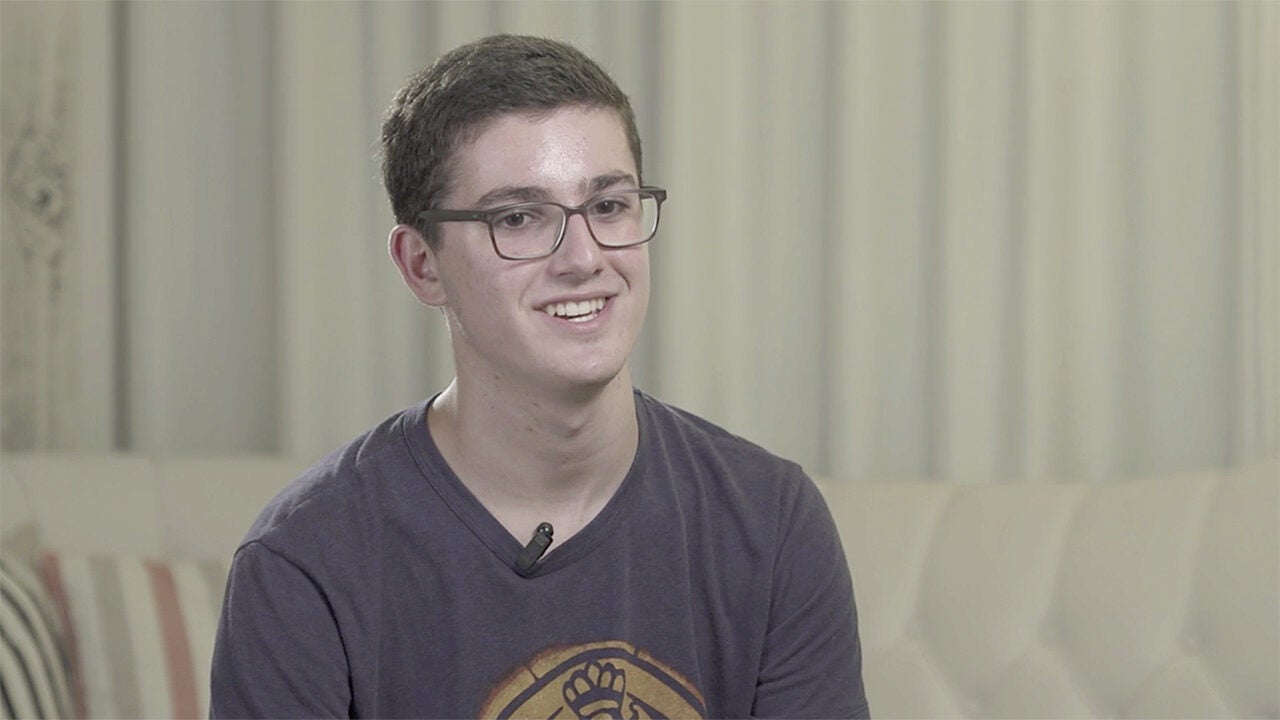 A person with short dark hair, wearing glasses and a t-shirt, sitting on a sofa and smiling.