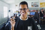 A person wearing glasses and a black shirt is smiling while holding a small electronic device in a classroom setting.