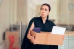 A person wearing a black outfit, holding a cardboard box with items, in an office setting. Their expression appears surprised or uncertain.
