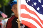 A child wearing a blue cap is partially obscured by a large American flag during a parade or public event.
