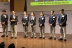 A group of students dressed in uniforms, standing on a stage during a presentation. The backdrop displays "Wharton High School Investment Competition" with company logos.