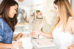 Two people in a jewelry store, one assisting the other in selecting a ring from a display case. A necklace is displayed on a bust in the background.