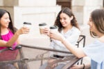 Three people sitting at an outdoor table, smiling, and toasting with coffee cups, representing friendship and socializing.