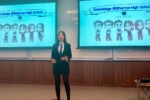 A person in a suit is giving a presentation in a classroom. Two screens display cartoon illustrations of people with the text "Knowledge @ Wharton High School" and "Pegasus."