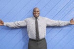 Person smiling with arms outstretched, wearing a checkered shirt and tie, standing against a textured blue wall.