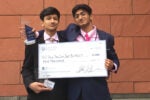 Two people in formal attire holding a large check for $5,000 and trophy awards, standing against a brick wall. The check is made out to "All You Can Eat Buffett."