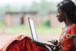 Person in a vibrant red dress sitting outdoors, using a laptop. The scene suggests connectivity and learning in a rural setting.
