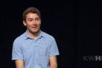 A person is sitting against a black background, wearing a light blue striped shirt. The letters "KWHS" are visible in the lower right corner.