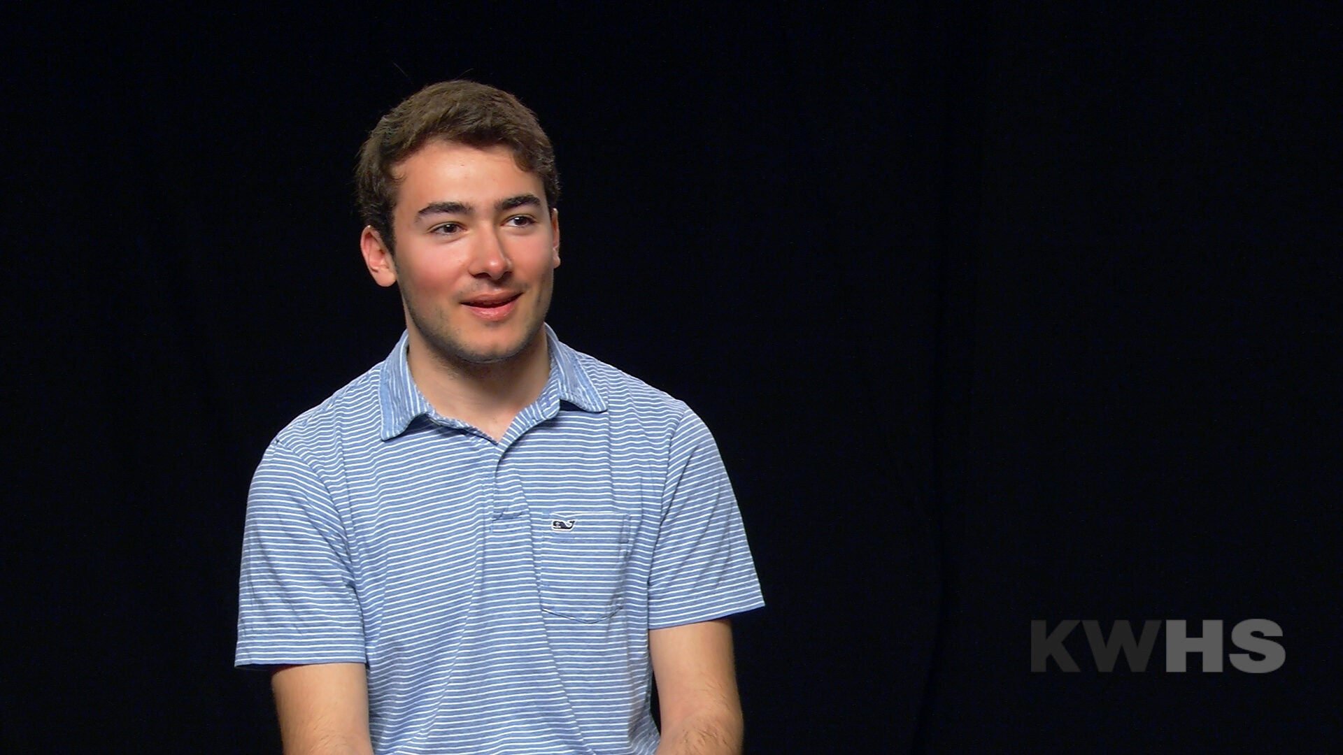 A person is sitting against a black background, wearing a light blue striped shirt. The letters "KWHS" are visible in the lower right corner.