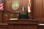 A person standing at a podium in a courtroom or legislative chamber, with the seal of the Florida State Senate in the background. The American and Florida state flags are visible.
