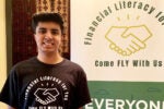 A person smiling beside a banner that says "Financial Literacy for You" and "Come FLY With Us" with an image of a handshake. The person is wearing a matching t-shirt.