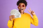 Person wearing a yellow sweater and round sunglasses, holding a fan of dollar bills in one hand and showing a peace sign with the other, against a purple background.