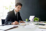 A person in a suit working at a desk with various papers, a laptop, a tablet, and a green mug, suggesting a business or office environment.