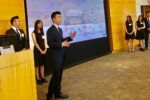 A group of six people in formal attire stand in a conference room giving a presentation about Alibaba. One person is speaking, while others stand near a podium and display screen.