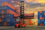 A forklift hoisting a red shipping container amidst stacks of red and blue containers under a dramatic sky.