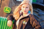 A person posing in front of a green tractor with a logo, wearing large turquoise earrings and a black jacket with "Heritage Family Farms" on it.