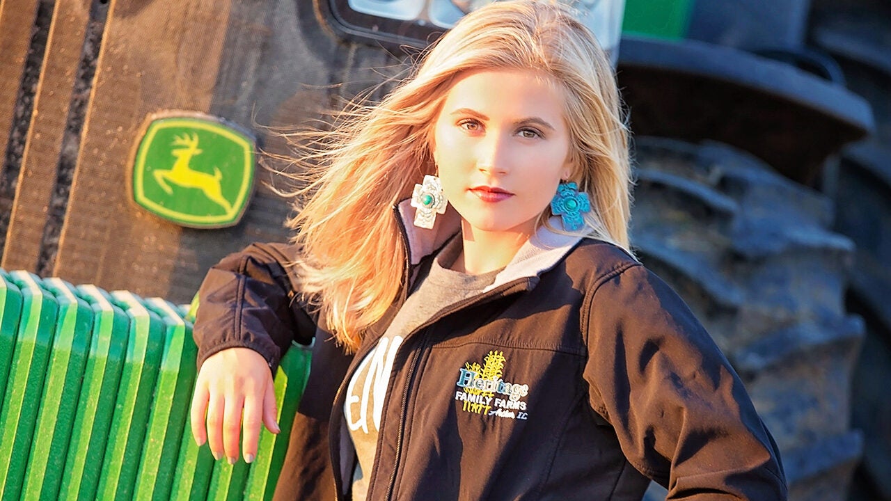 A person posing in front of a green tractor with a logo, wearing large turquoise earrings and a black jacket with "Heritage Family Farms" on it.