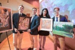 Four individuals holding framed artworks, smiling in a gallery setting. The art includes a surreal portrait, a landscape, and abstract designs.