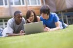 Three people lying on grass, looking at a laptop together, suggesting a collaborative or social activity in an outdoor setting.