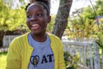 A smiling person wearing a yellow cardigan and graphic T-shirt stands outdoors near a white picket fence, surrounded by greenery.