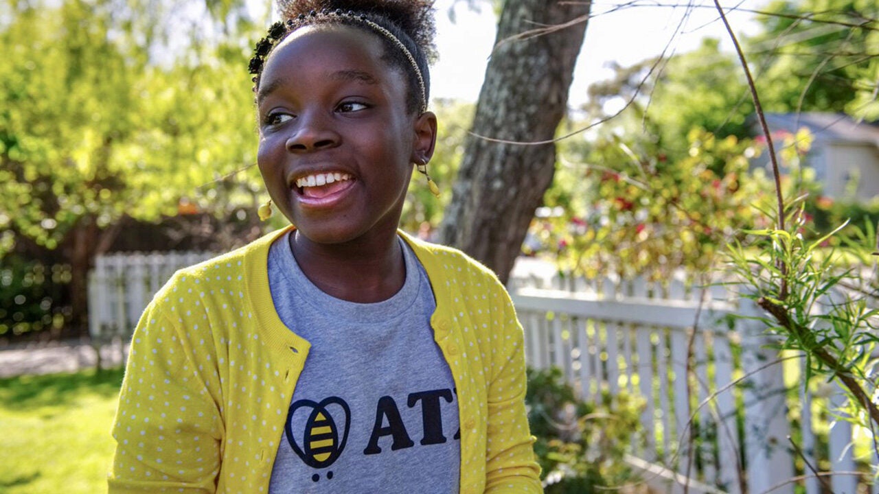 A smiling person wearing a yellow cardigan and graphic T-shirt stands outdoors near a white picket fence, surrounded by greenery.