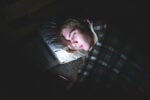 A person lying in bed under a plaid blanket, illuminated by the light from a smartphone they are holding, in a dark room.