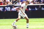 Soccer player in a white uniform dribbling a ball on a field, with spectators in the background.