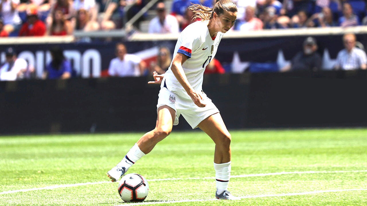 Soccer player in a white uniform dribbling a ball on a field, with spectators in the background.