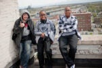 Three people standing on a rooftop with an urban landscape in the background. One is giving a thumbs-up, one holds headphones and a camera, and the third stands with arms crossed.