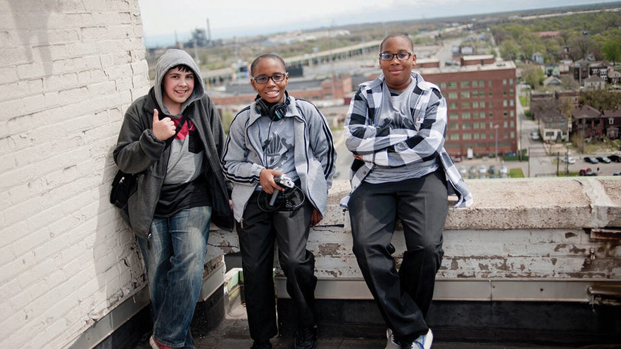 Three people standing on a rooftop with an urban landscape in the background. One is giving a thumbs-up, one holds headphones and a camera, and the third stands with arms crossed.